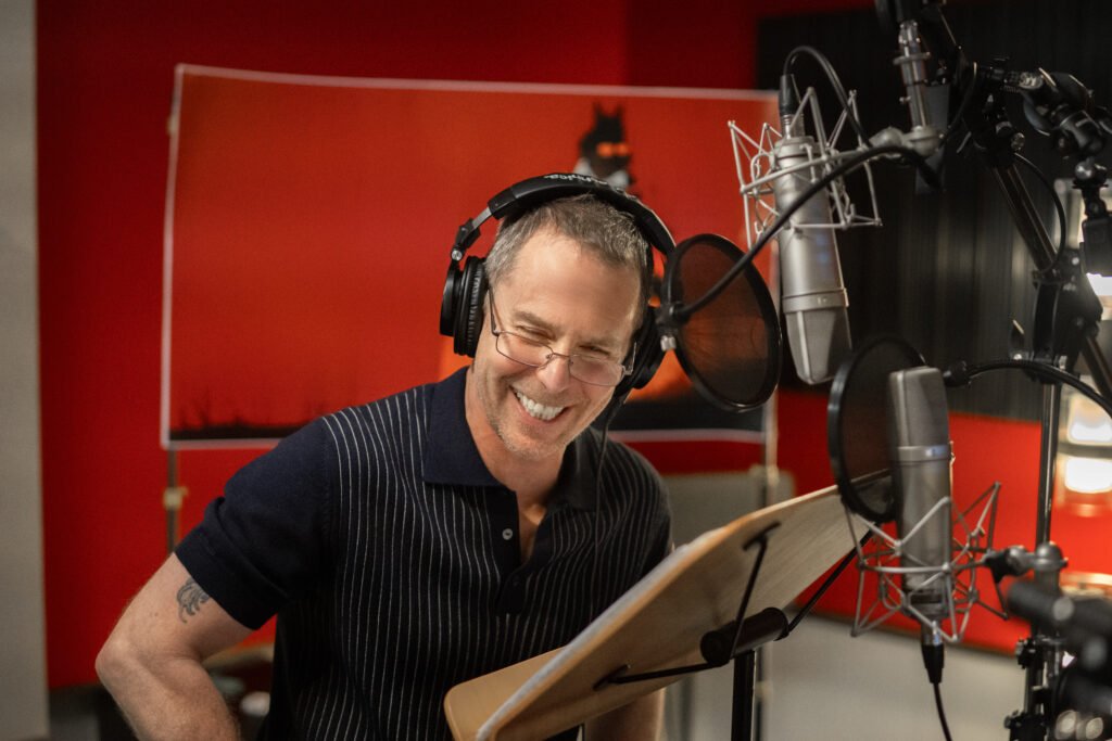 alt="Behind‑the‑scenes photo of Sam Rockwell in a recording booth wearing headphones and reading from a script while voicing Wolf for The Bad Guys 2."
