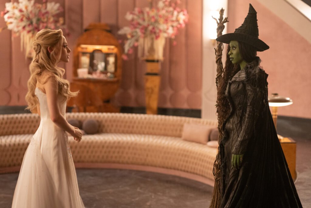 alt="Scene from 'Wicked for Good' showing Glinda in a flowing white gown and Elphaba in a dark witch costume with green skin standing together in an ornate, warmly lit room with a curved sofa and floral arrangements."
