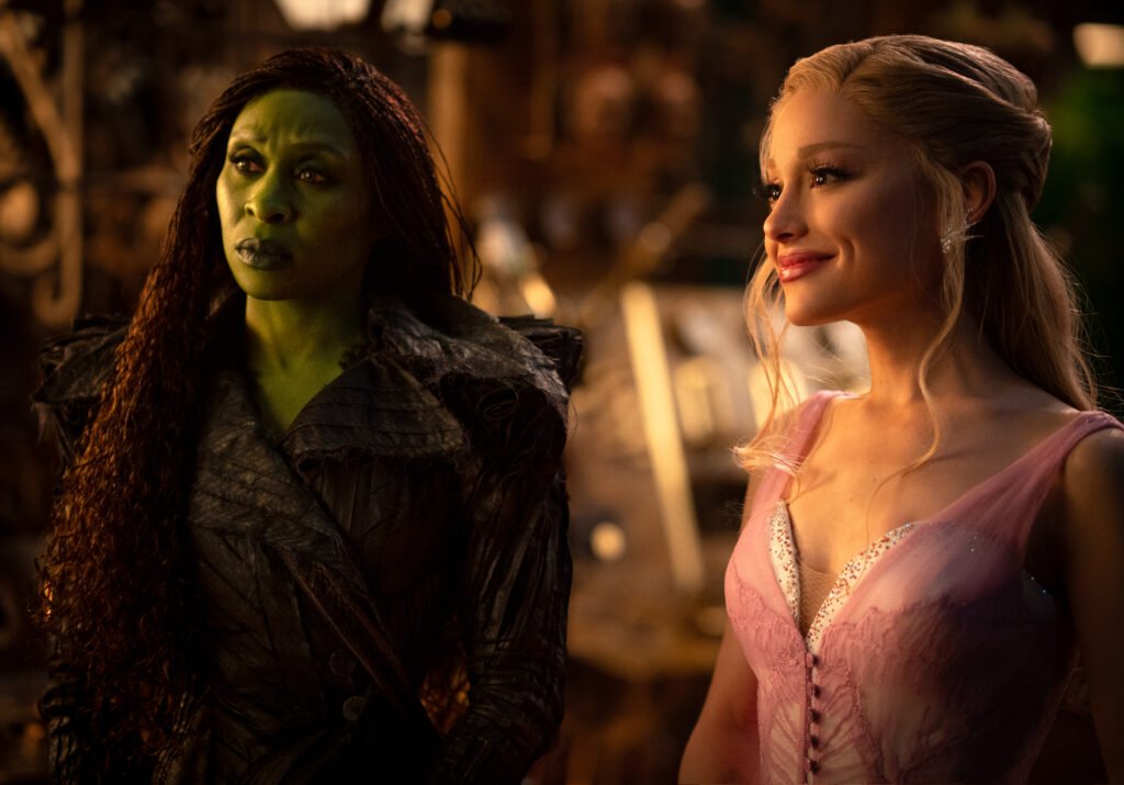 alt="Scene from 'Wicked for Good' showing Elphaba with green skin in a dark textured outfit standing beside Glinda in a pale pink lace dress, both framed in a warmly lit, ornate room that enhances the film’s magical atmosphere."
