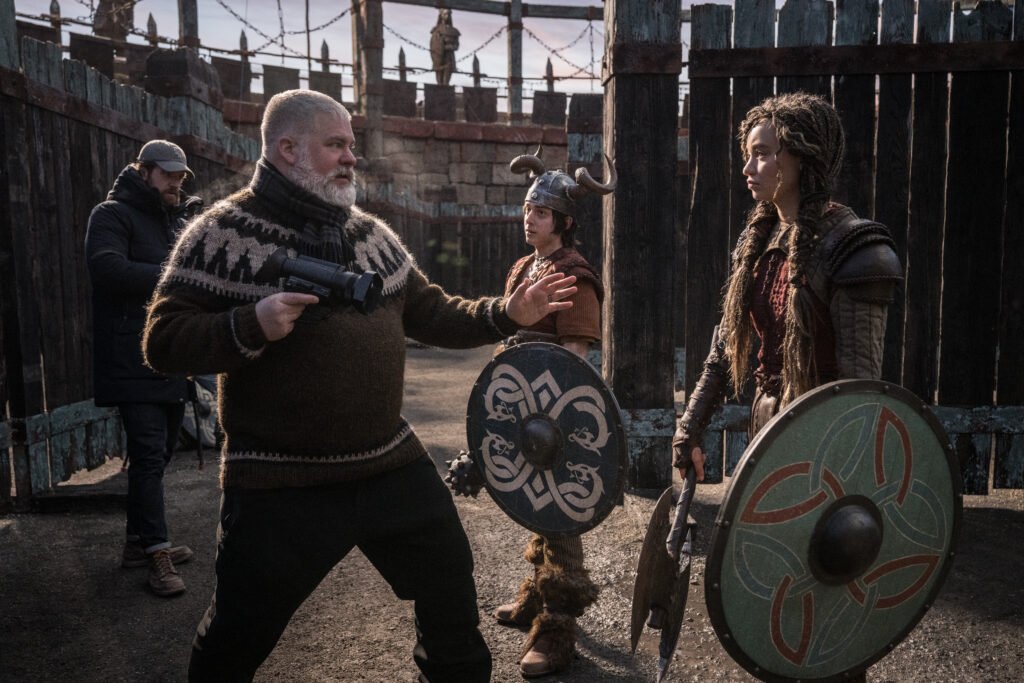 alt="Behind‑the‑scenes photo from the live‑action 'How to Train Your Dragon' showing writer‑director Dean DeBlois with actors Gabriel Howell and Nico Parker in Viking‑style costumes on a rustic village set."
