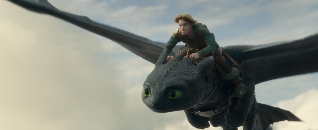 alt="Scene from the live‑action 'How to Train Your Dragon' showing Toothless the Night Fury flying through a cloudy sky with Hiccup, played by Mason Thames, riding on his back."
