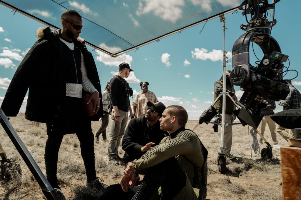 alt="Behind‑the‑scenes image from 'HIM (2025)' showing Marlon Wayans, director Justin Tipping, and Tyriq Withers talking together on an outdoor set with dry grass, a large camera rig, and a partly cloudy sky."
