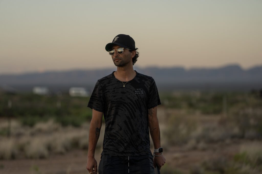 alt="Behind‑the‑scenes image from 'HIM (2025)' showing director Justin Tipping standing outdoors in a desert landscape at dusk, wearing a cap, sunglasses, and a dark patterned shirt."
