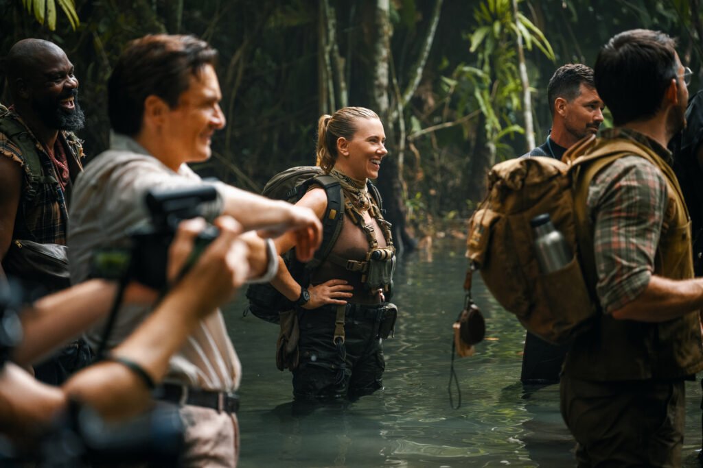 alt="Behind‑the‑scenes photo from 'Jurassic World: Rebirth' showing Scarlett Johansson and other cast members standing in shallow jungle water wearing tactical gear during a scene setup."
