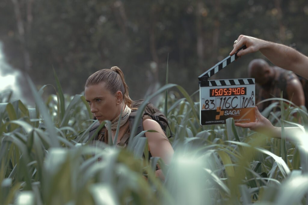 alt="Behind‑the‑scenes photo from 'Jurassic World: Rebirth' showing Scarlett Johansson as Zora Bennett crouched in tall grass with a clapperboard visible in the foreground."

