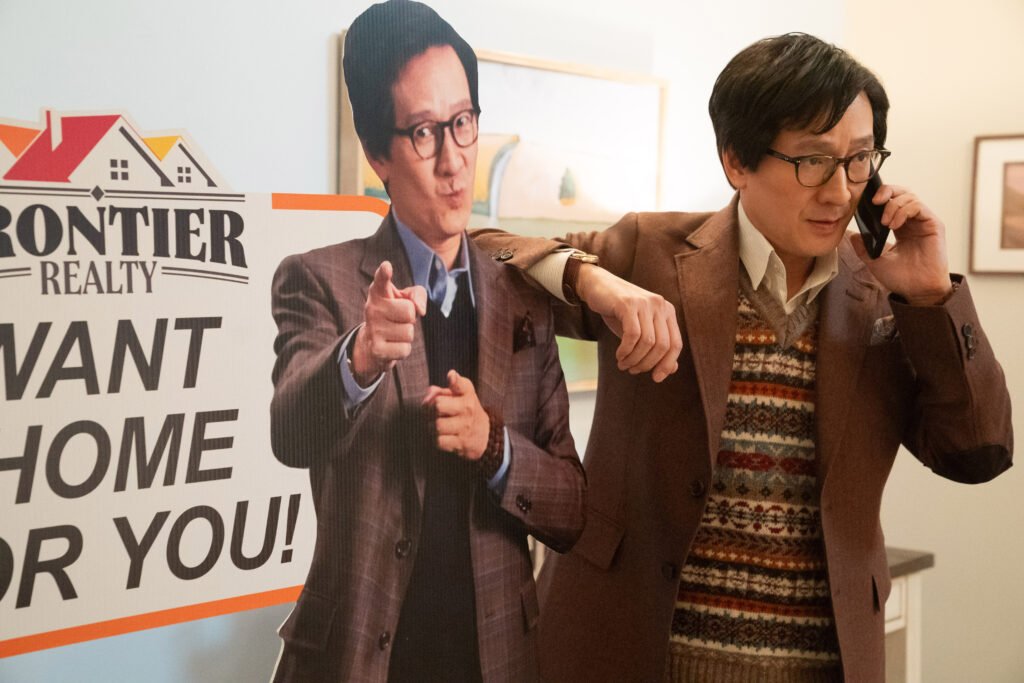 alt="Scene from 'Love Hurts' showing Ke Huy Quan as Marvin Gable standing beside a life‑size real‑estate cutout of himself while talking on a phone."
