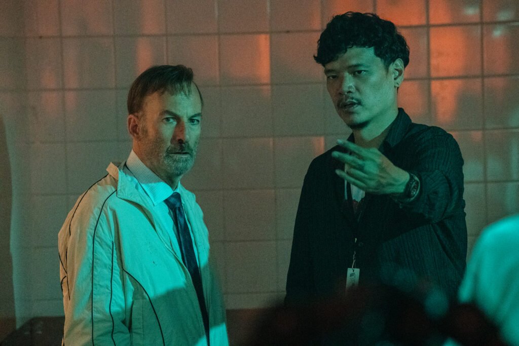 alt="Behind‑the‑scenes photo from 'Nobody 2' showing Bob Odenkirk standing beside director Timo Tjahjanto in a dimly lit tiled room with green and orange lighting."
