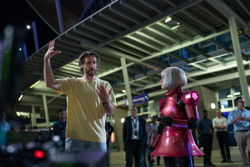 alt="Behind‑the‑scenes photo from 'M3GAN 2.0' showing director Gerard Johnstone speaking with a performer in a metallic pink robotic costume on a busy set."
