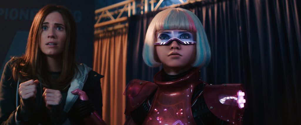 alt="Behind‑the‑scenes image from 'M3GAN 2.0' showing Gemma, played by Allison Williams, standing beside the futuristic robot M3GAN in a stage‑lit setting with curtains and equipment in the background."
