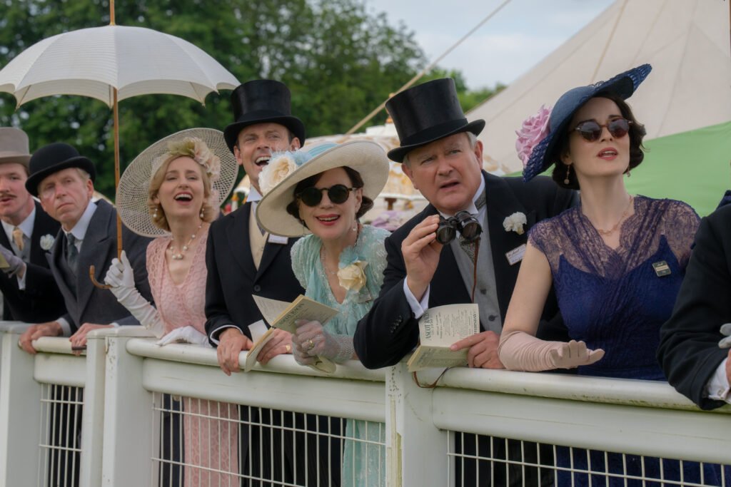 alt="Scene from 'Downton Abbey: The Grand Finale' showing Lady Edith, Bertie Hexham, Cora Grantham, Robert Grantham, and Lady Mary standing behind a white fence at an outdoor formal event, dressed in elegant attire with hats, gloves, and programs."