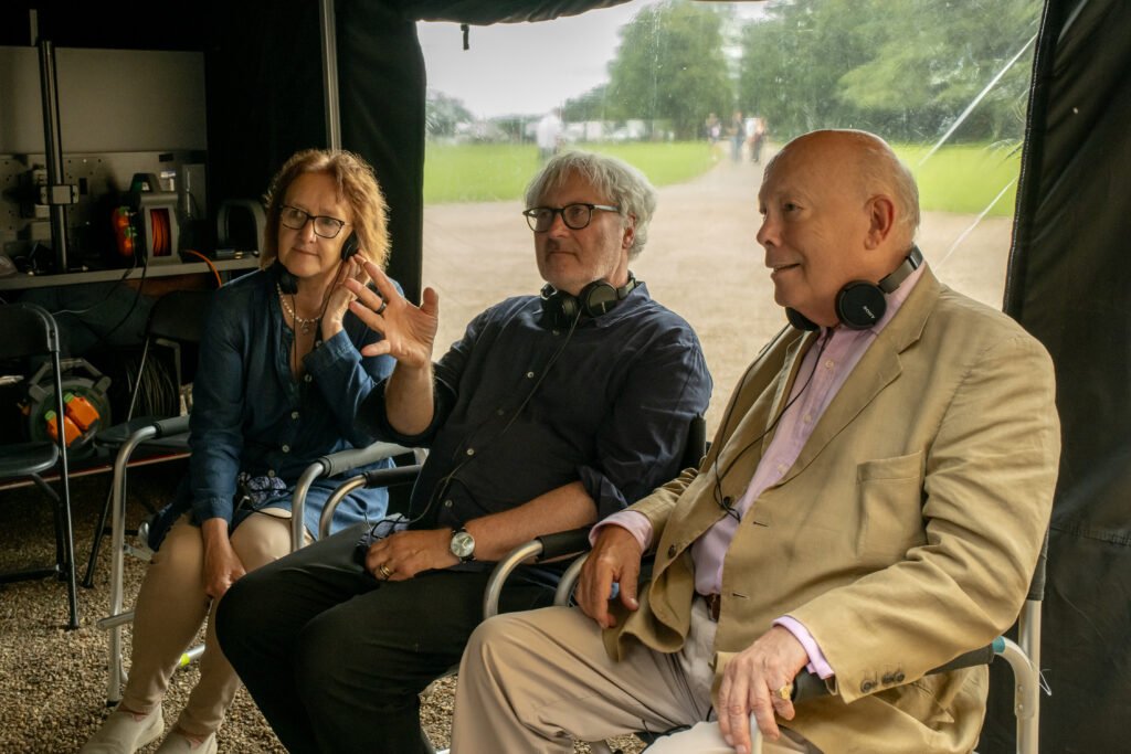 alt="Behind‑the‑scenes image from 'Downton Abbey: The Grand Finale' showing producer Liz Trubridge, director Simon Curtis, and writer Julian Fellowes seated under a tent on set, discussing the production with equipment visible behind them."