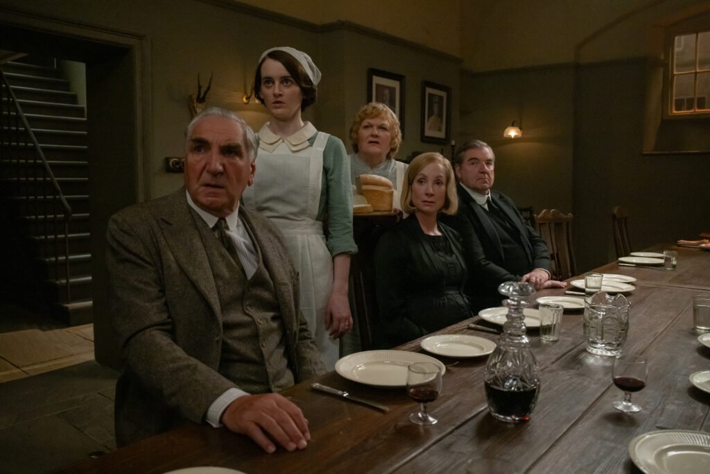 alt="Scene from 'Downton Abbey: The Grand Finale' showing Mr. Carson, Daisy Parker, Mrs. Patmore, Anna Bates, and Mr. Bates seated around a dimly lit wooden dining table set with plates, cutlery, and glasses of red wine."