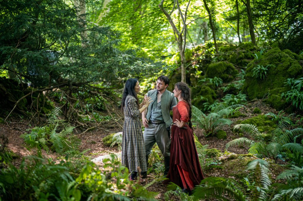 alt="Behind‑the‑scenes photo from the film 'Hamnet' showing director Chloé Zhao standing in a lush green forest with actors Paul Mescal and Jessie Buckley, all dressed in period costumes as they discuss a scene among dense ferns and moss‑covered trees."
