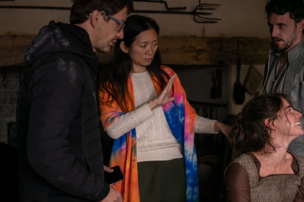 alt="Behind‑the‑scenes photo from the film showing director Chloé Zhao speaking with director of photography Lukasz Zal and actors Jessie Buckley and Paul Mescal inside a rustic, dimly lit set as they discuss a scene."
