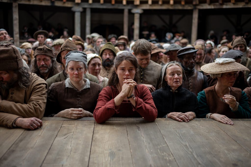 Alt = "Scene from Chloé Zhao’s Hamnet showing Jessie Buckley as Agnes and Joe Alwyn as Bartholomew surrounded by solemn townspeople, used in coverage of the film’s presence at the Oscars 2026."