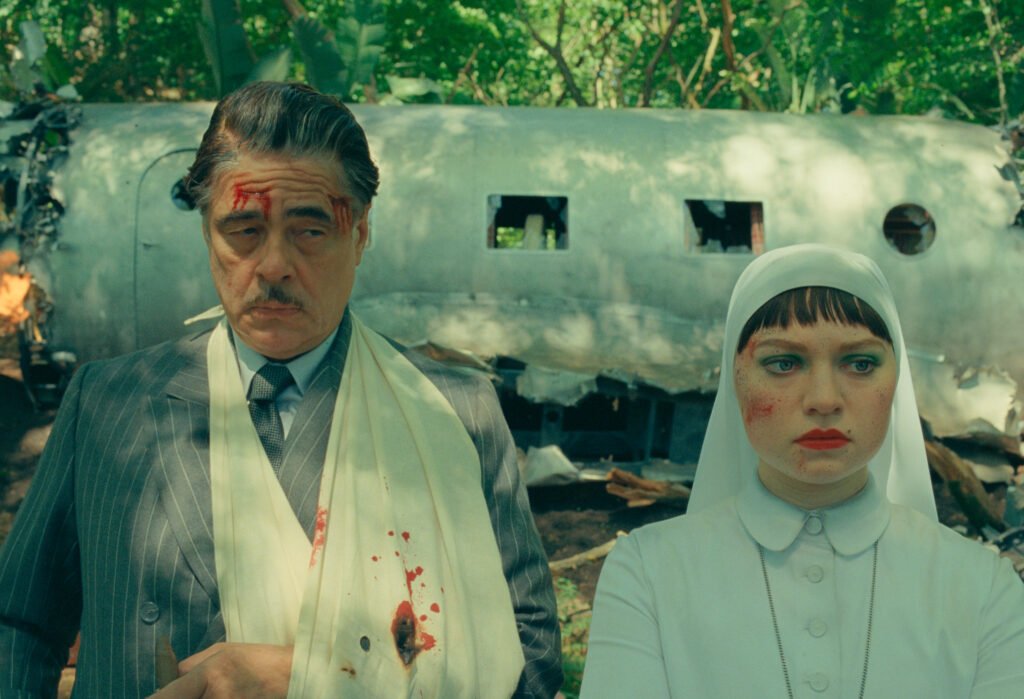 alt="Scene from 'The Phoenician Scheme' showing two injured characters standing before a crashed airplane in a forest, including a man in a pinstripe suit and a woman in a white habit."