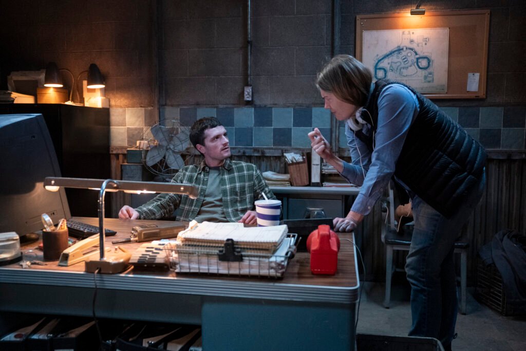 alt="Behind‑the‑scenes photo from 'Five Nights at Freddy’s 2' showing Josh Hutcherson seated at a cluttered desk in a dim control‑room set while director Emma Tammi stands beside him giving direction, with blue‑tiled walls, a fan, and a bulletin board in the background."
