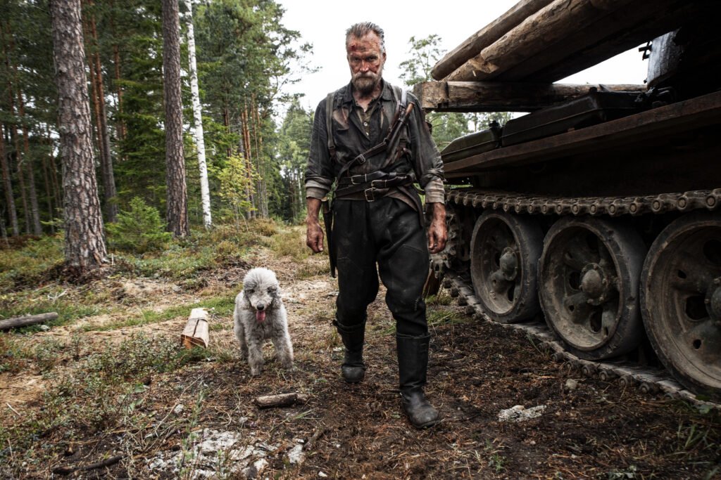 alt="Promotional still from the film 'Sisu: Road to Revenge' showing a rugged, dirt‑covered man walking through a forest beside a large military tank with a dog at his side, creating an intense, war‑torn atmosphere."
