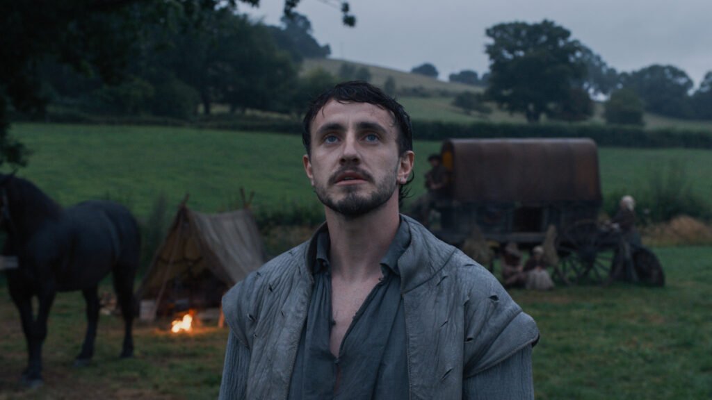 alt="Scene from the film 'Hamnet' showing Paul Mescal in period costume standing in a misty rural field beside a horse, with a tent, wagon, and campfire behind him, creating a somber, atmospheric setting."
