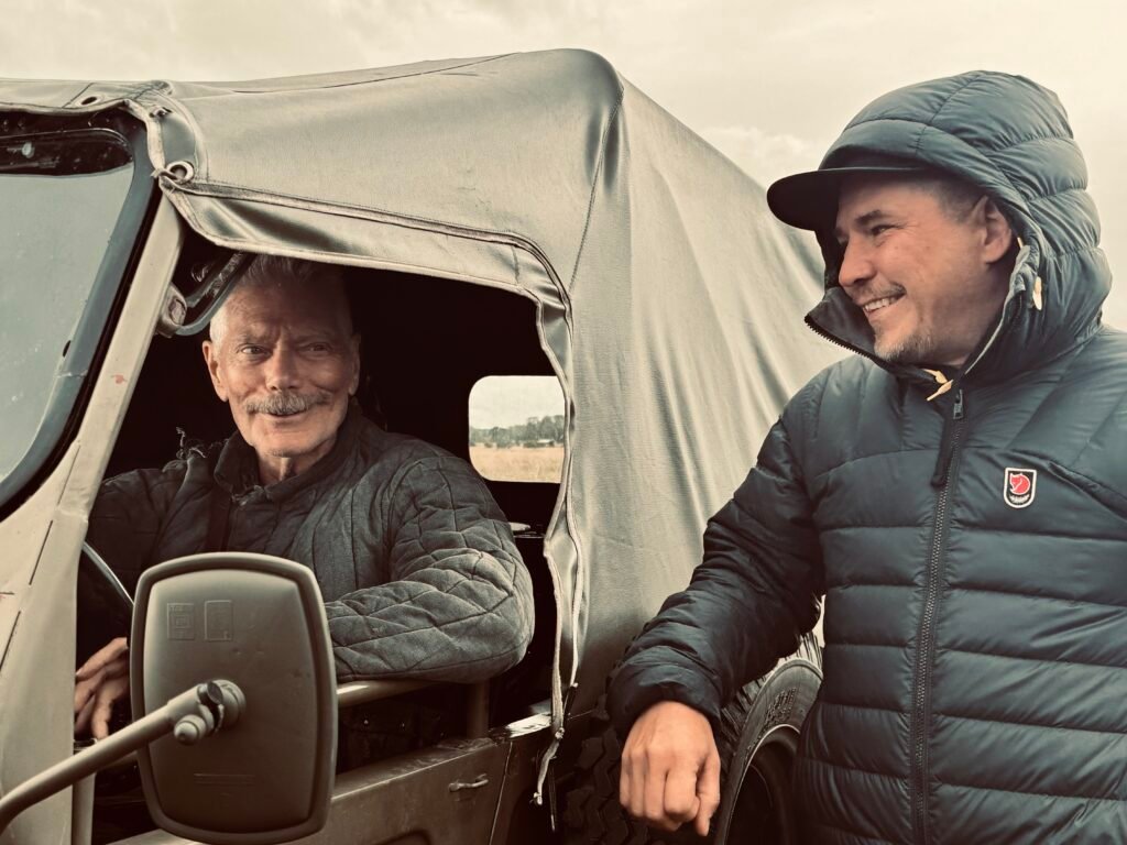 alt="Behind‑the‑scenes photo from the film 'Sisu: Road to Revenge' showing an actor seated in the driver’s seat of a military-style vehicle while the director stands beside it, both wearing heavy quilted outerwear in a cold outdoor location with fields in the background."
