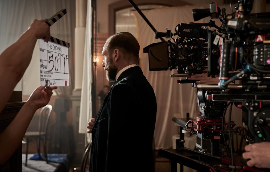 alt="Behind‑the‑scenes photo from the film 'The Choral' showing actor Ralph Fiennes in vintage costume standing on set as a clapperboard is held in front of him, with camera equipment and crew preparing a shot inside a staged period environment."
