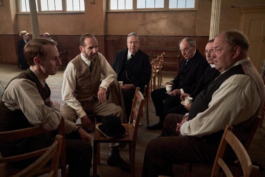 alt="Behind‑the‑scenes photo from the film 'The Choral' showing seven men in period clothing seated in a circle on wooden chairs in a bright room with tall windows and a wooden floor, sharing tea during a break on set."
