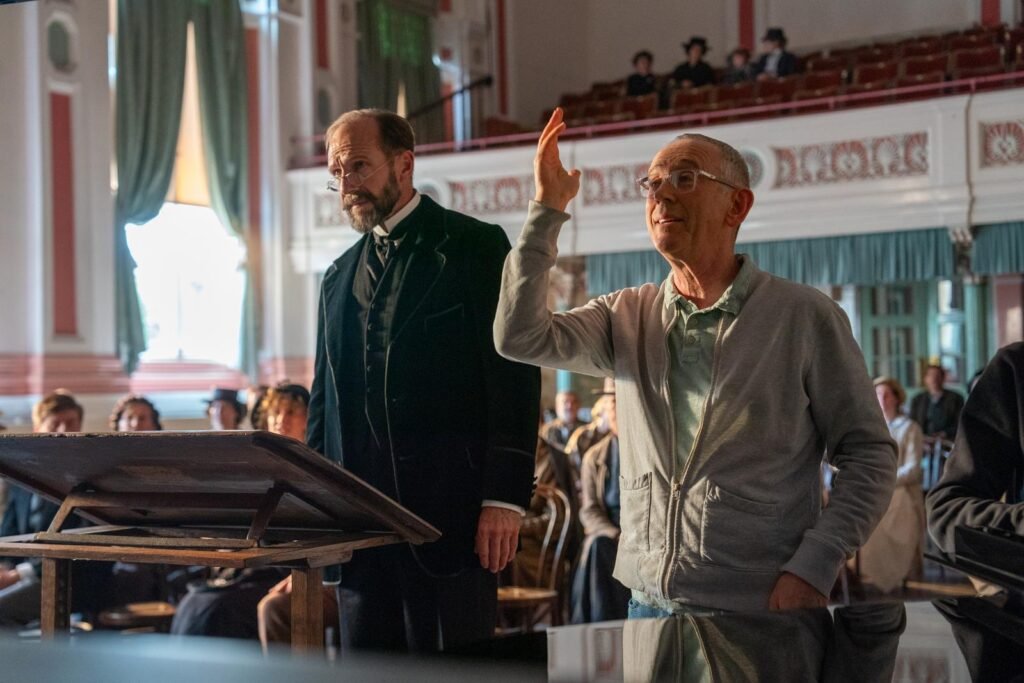 alt="Behind‑the‑scenes photo from the film 'The Choral' showing the actor Ralph Fiennes in period costume standing beside a music stand while the director Nicholas Hytner gestures instructions in a grand hall filled with seated extras in vintage clothing."

