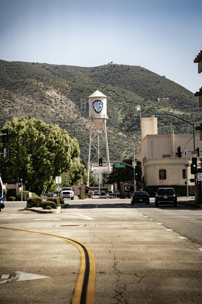 alt="Street view leading toward the Warner Bros. water tower with the WB logo, framed by trees, traffic lights, and passing cars, used to illustrate the headline 'Paramount Wins Warner Netflix Battle.'"
