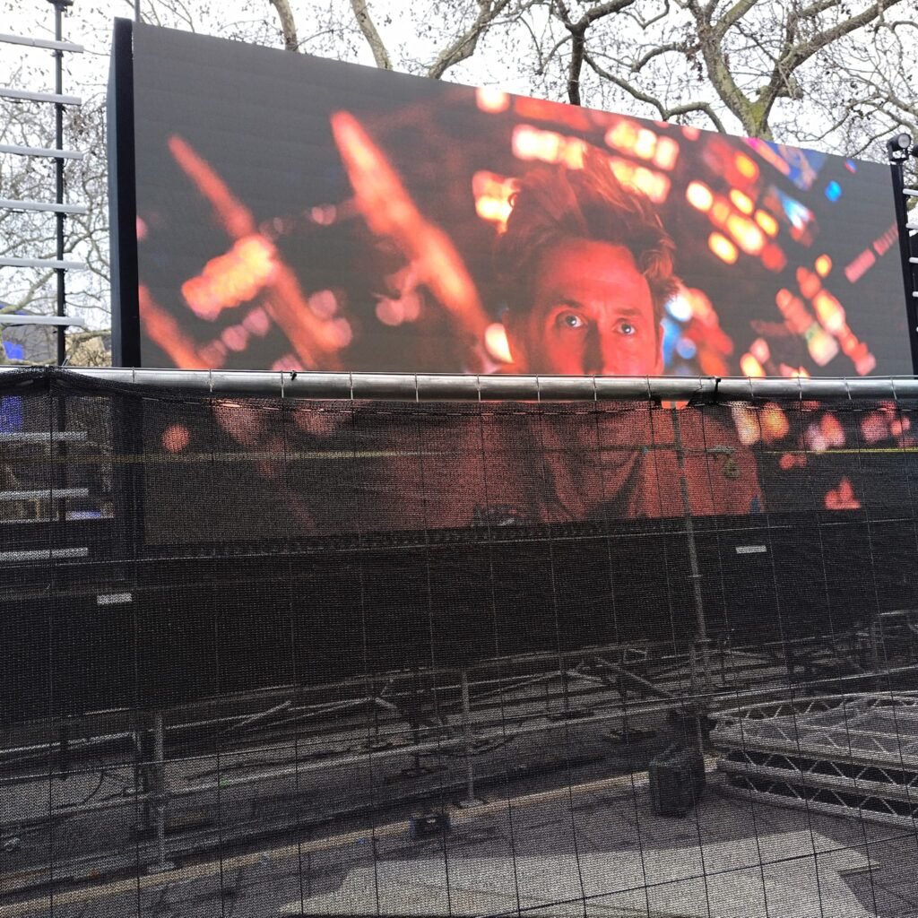 Alt text:  
Outdoor setup for the Project Hail Mary London premiere, featuring a large LED screen displaying a close‑up film scene with metal framing and mesh barriers in front.