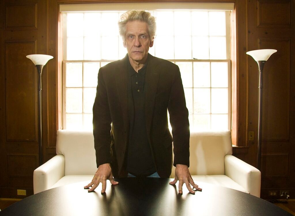 Alt = David Cronenberg stands behind a black table in a warmly lit room, leaning forward with both hands on the tabletop. He wears a dark blazer and shirt, framed by a large multi‑pane window, white armchairs, and tall floor lamps that create a dramatic, authoritative atmosphere. David Cronenberg’s birthday