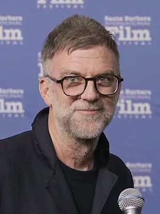 Alt = Alt text:  "Paul Thomas Anderson speaking into a microphone in front of a blue Santa Barbara International Film Festival backdrop, featured in coverage related to Paul Thomas Anderson and the Oscars 2026