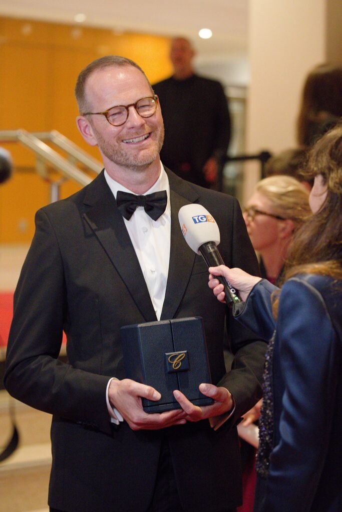 Alt = "Director Joachim Trier, dressed in a black tuxedo, smiles while holding a dark award box with a gold emblem as he’s interviewed on the red carpet by Italian news channel TG5 during an event."