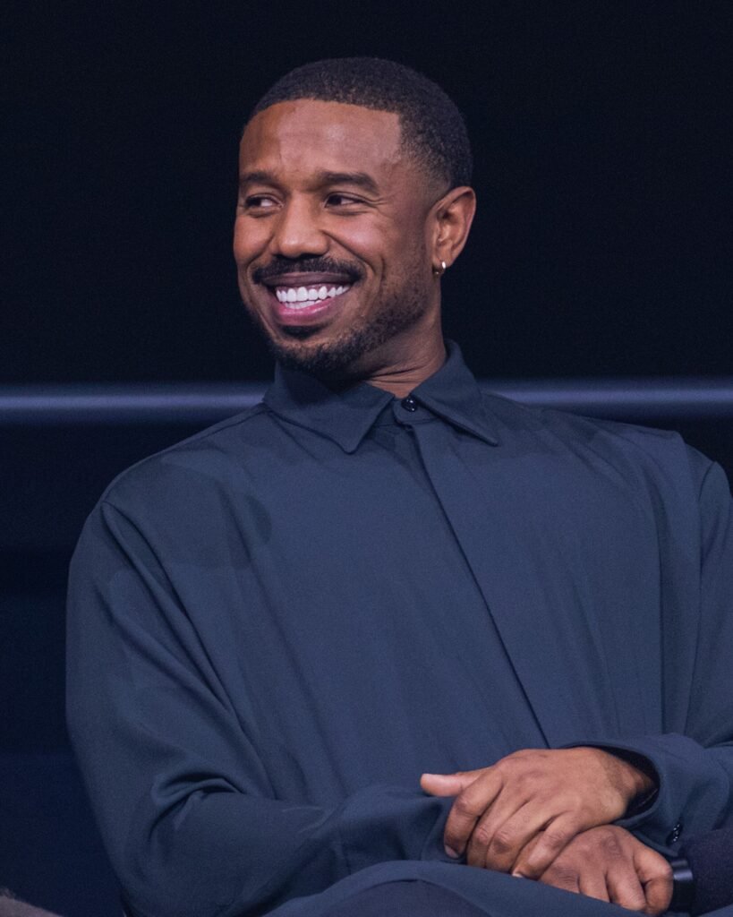 Alt ="Michael B. Jordan sits smiling in a dark button‑up shirt against a shadowed backdrop during an interview moment connected to the Oscars 2026 season."