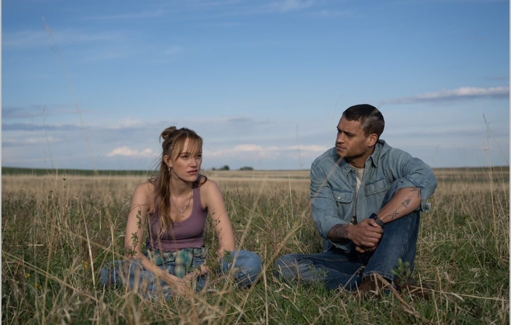 Alt = Maika Monroe and Tyriq Withers sit together in a grassy field under a bright sky, talking quietly in a calm, reflective moment from the romantic drama Reminders of Him.