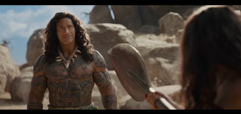 Alt =Alt text:  
Live‑action “Moana” 2026 scene featuring Dwayne Johnson as Maui, standing in a rocky sunlit landscape with his Polynesian tattoos visible as he faces someone holding a curved weapon, used in a Moana live action trailer discussion.