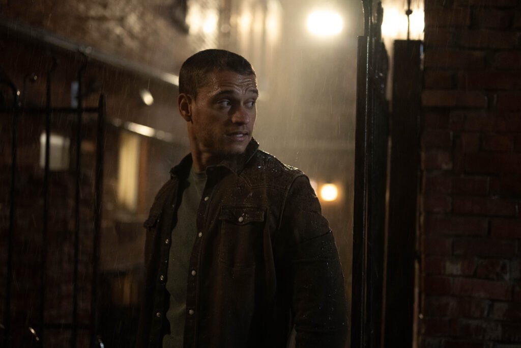 Alt =  A tense, rain‑lit alley scene featuring Tyriq Withers as Ledger Ward in Reminders of Him, standing in a dark jacket and looking off to the side as warm lights glow behind him.