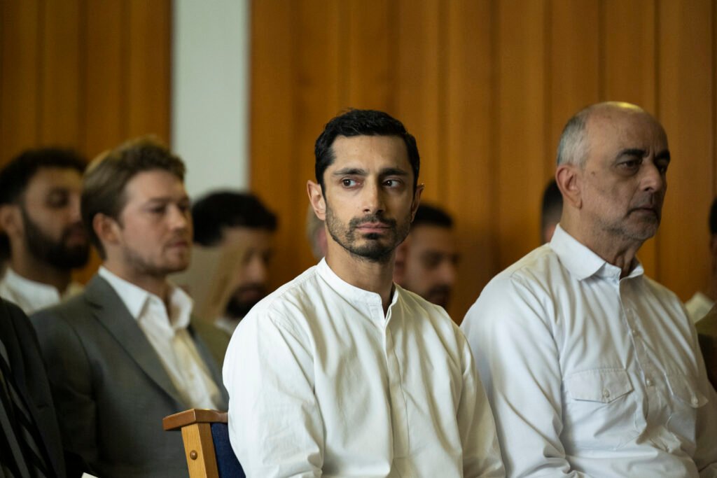 Alt=  Riz Ahmed sits at the center of a formal gathering beside Joe Alwyn and Art Malik, surrounded by men in suits and traditional attire in a solemn, wood‑paneled setting Hamlet 2025