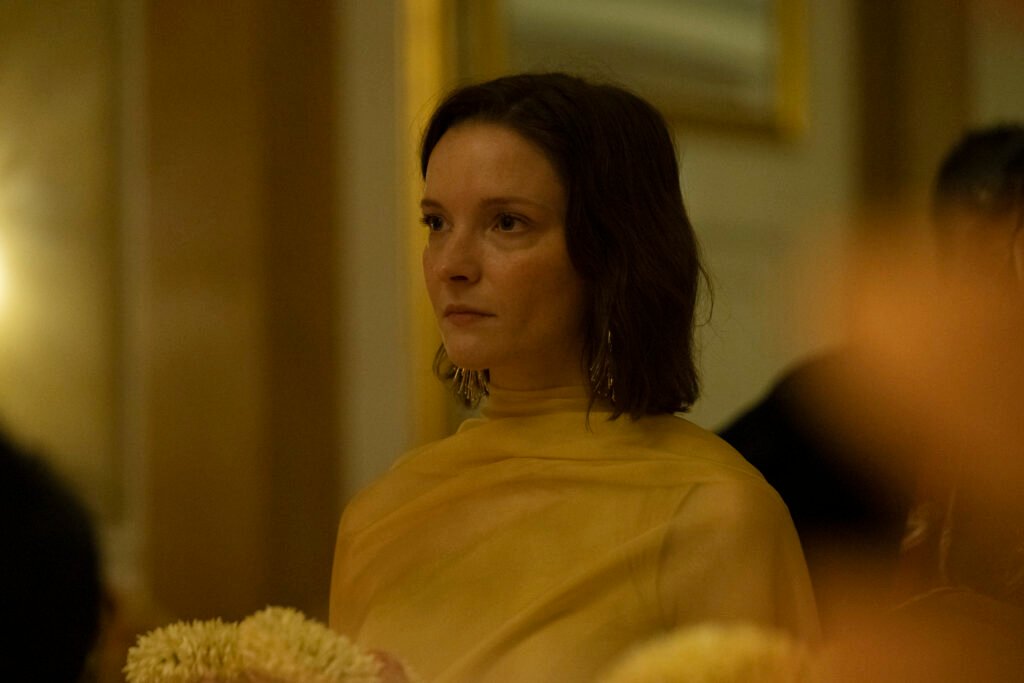alt= Morfydd Clark dressed in a yellow garment during an emotional moment in a softly lit scene from Hamlet 2025 with blurred flowers in the foreground.