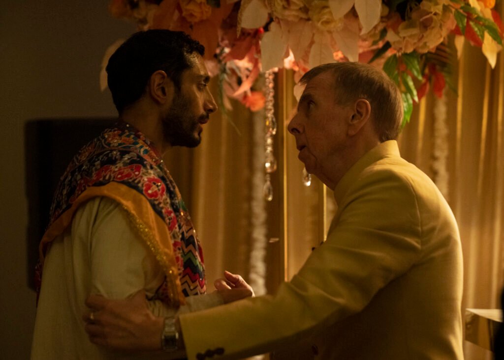 Alt=Riz Ahmed and Timothy Spall stand closely together under a canopy decorated with hanging flowers and crystals in an intense moment from a dramatic scene, with warm lighting enhancing the emotional atmosphere Hamlet 2025