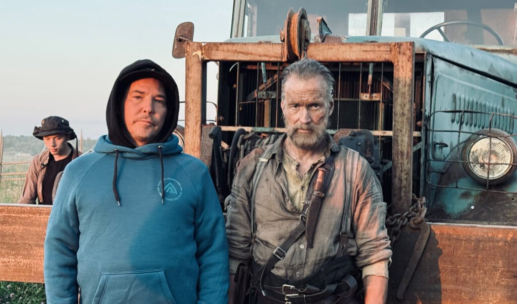 Alt text:  
Director Jalmari Helander and actor Jorma Tommila standing beside a large rusted industrial vehicle on the set of SISU 2: ROAD TO REVENGE, joined by another crew member in an outdoor filming location.