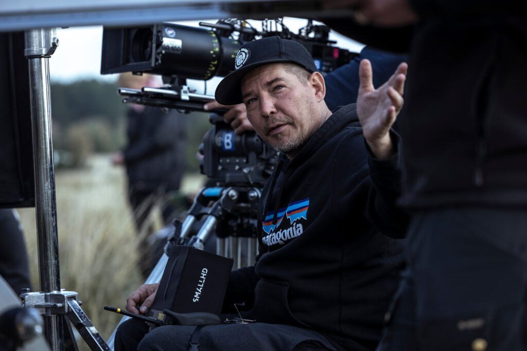 Alt text:  
Film director Jalmari Helander wearing a black hoodie and cap reviewing notes beside a professional camera on set, surrounded by crew and equipment during an outdoor production.
