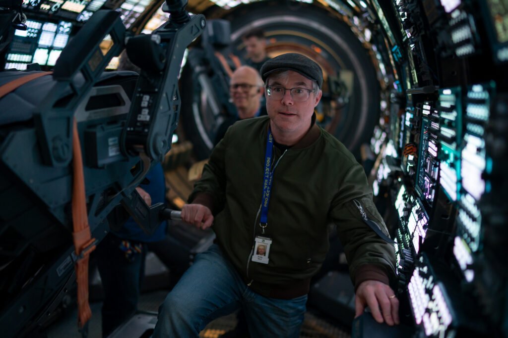 Alt = Alt attribute:  
Author Andy Weir stands inside a detailed spacecraft‑style set surrounded by futuristic control panels during the production of Project Hail Mary, shown here to accompany a Project Hail Mary movie review.