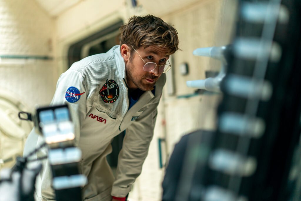 Alt = Ryan Gosling in a white astronaut suit leans forward inside a spacecraft‑style set in a promotional still from Project Hail Mary, shown here to accompany a Project Hail Mary movie review.