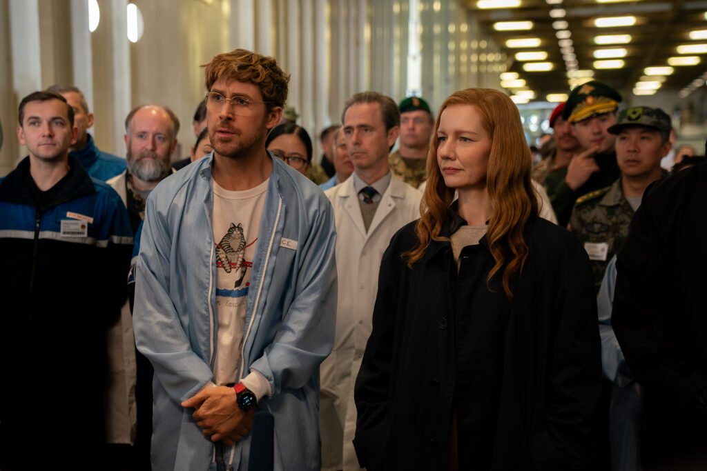 Alt = Ryan Gosling as Ryland Grace stands beside Sandra Hüller as Eva Stratt in a brightly lit corridor with scientists and military personnel in the background, shown here to accompany a Project Hail Mary movie review.