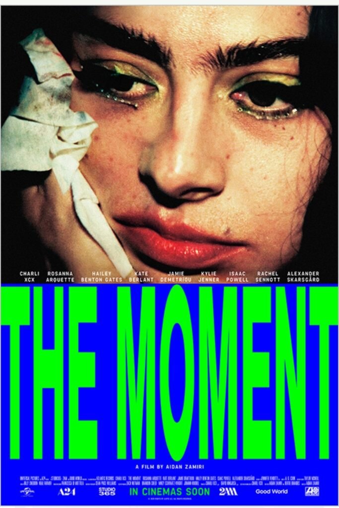 Alt text:  
Close-up of Charlie xcx with bold green eye makeup holding a tissue to her face, with the title “The Moment” in large green letters below